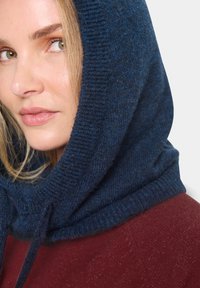 Căciulă tricotată de culoare albastru marin, cu un finisaj texturat, având un detaliu cu șnur. Modelul poartă un top burgund, punând în evidență designul stratificat al căciulii.