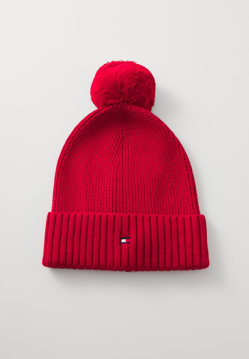 Rød strikket lue med ribbestrikket kant, toppet med en luftig pom-pom. Har et lite logo-merke i blått, hvitt og rødt på kanten.