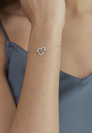 Silberarmband mit einem herzförmigen Anhänger, der mit klaren Steinen besetzt ist, an einer zarten Kette befestigt, am Handgelenk getragen.