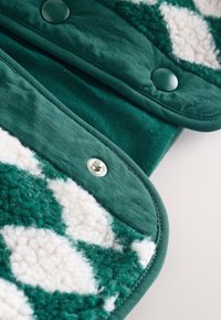 Tessuto verde caratterizzato da un materiale morbido e soffice con motivi a diamante bianchi. Comprende un tessuto liscio, chiusura a scatto e trame contrastanti.