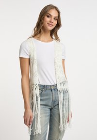Gilet bianco all'uncinetto aperto davanti con dettagli in frange, realizzato in filato leggero. Indossato sopra una maglietta bianca semplice e jeans azzurri chiari.