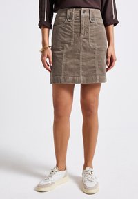 Kvinna klädd i en brun corduroy-minikjol med framfickor, vita sneakers och en brun topp med uppvikt ärm.