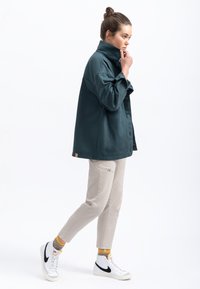Giacca camicia oversized color turchese con colletto alto e chiusura con bottoni, abbinata a pantaloni beige con un motivo ricamato e sneakers bianche.
