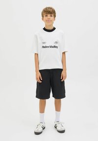 T-shirt blanche à manches courtes avec impression noire "Palm Valley", associée à un short cargo noir. Des chaussettes blanches et des baskets noir/blanc complètent la tenue.
