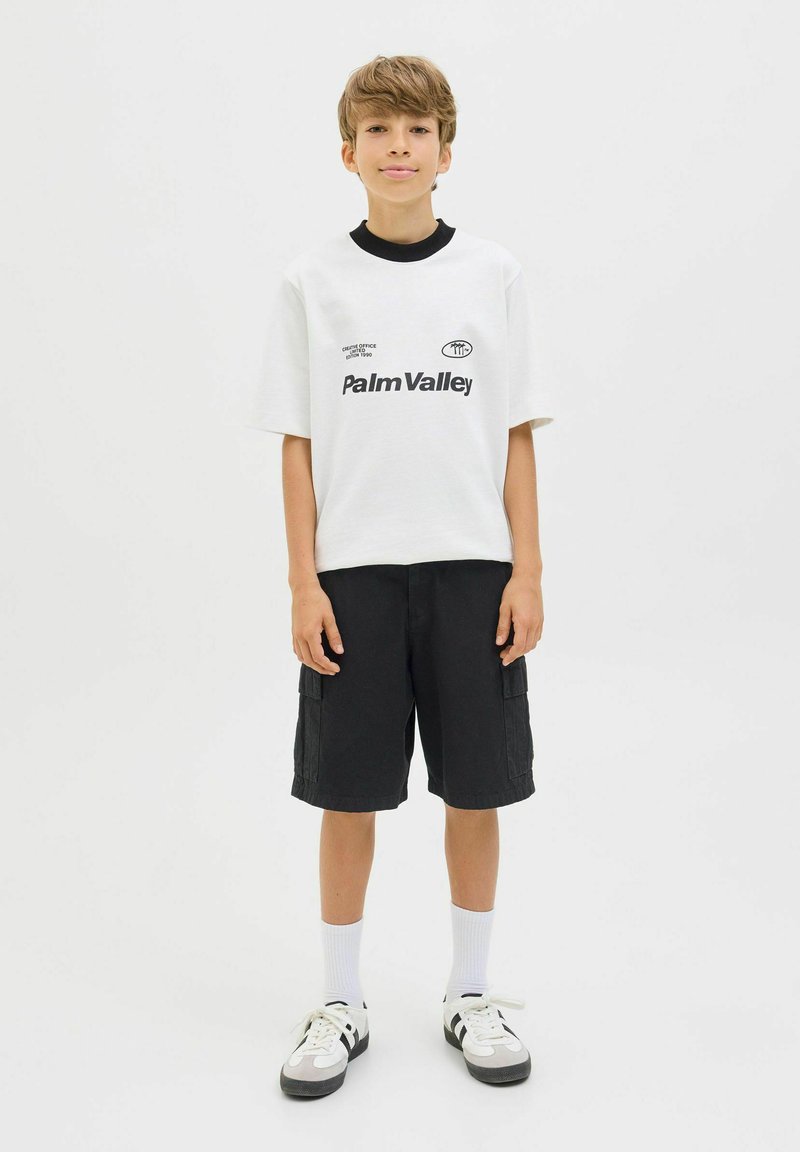 T-shirt blanche à manches courtes avec impression noire "Palm Valley", associée à un short cargo noir. Des chaussettes blanches et des baskets noir/blanc complètent la tenue.