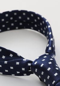 Bandeau en tissu navy avec un motif carré blanc, doté d'un design noué. Fabriqué en matériau lisse et brillant avec une structure flexible.