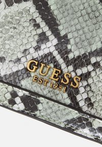 Texturovaná peněženka s hadím vzorem v odstínech zelené a černé, se zlatým logem "GUESS EST 1981" a hladkým, lesklým povrchem.