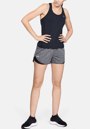 Donna in piedi con le mani sui fianchi, indossa una canotta nera senza maniche, pantaloncini sportivi grigi e scarpe da corsa nere su sfondo bianco.
