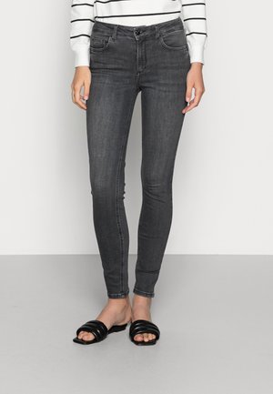 Kvinde iført mørkegrå skinny jeans, sorte åbne tåsandaler og en hvid langærmet top med sorte horisontale striber.