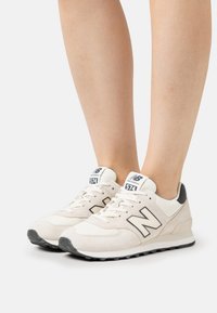 Białe zamszowe trampki z czarnymi akcentami, z wyraźnym logo "N", sznurowanym designem, wyściełanym kołnierzem i teksturowaną podeszwą.