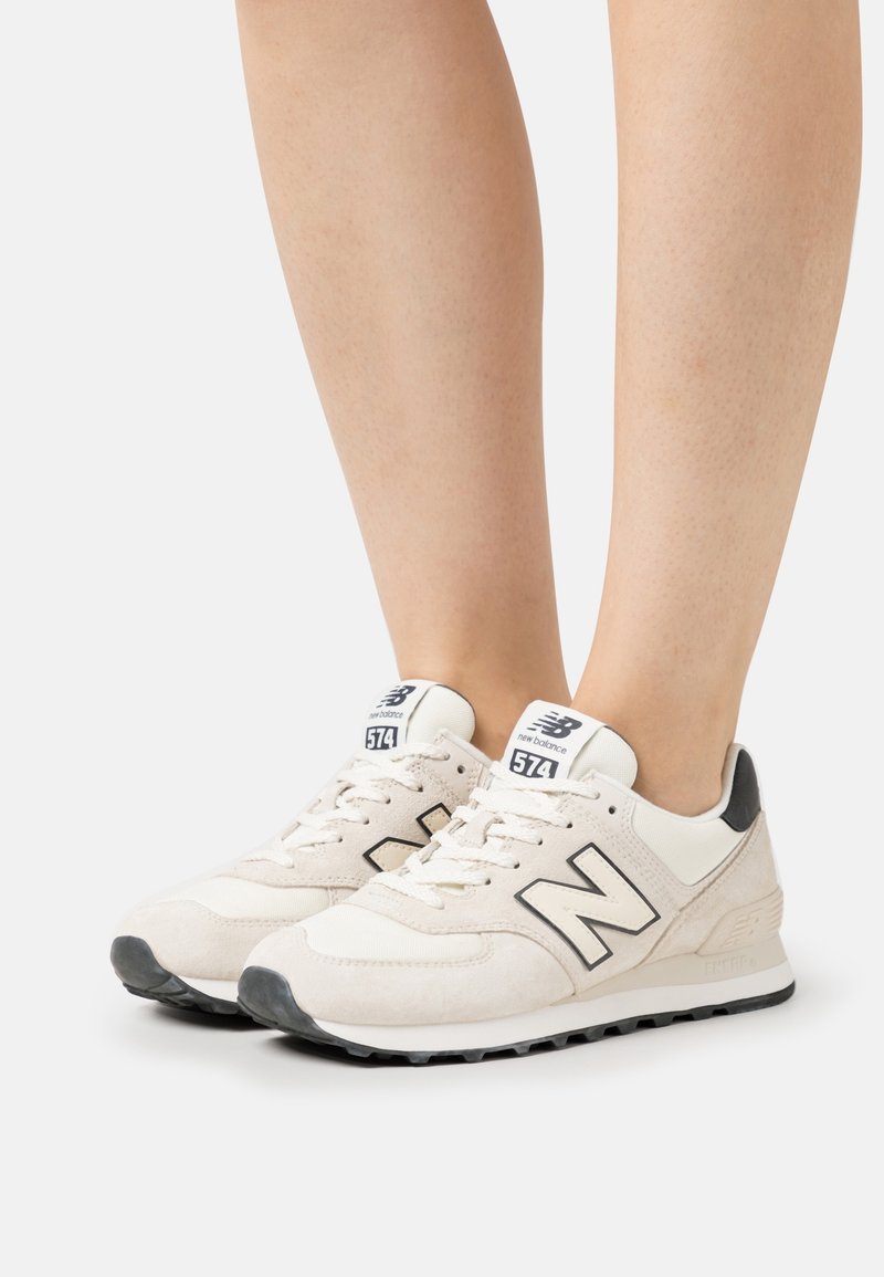 Białe zamszowe trampki z czarnymi akcentami, z wyraźnym logo "N", sznurowanym designem, wyściełanym kołnierzem i teksturowaną podeszwą.