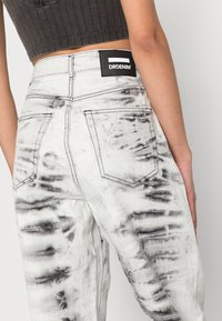 Jeans de ganga brancos com um padrão de tie-dye preto, cinco bolsos e uma etiqueta preta com marca na cintura.