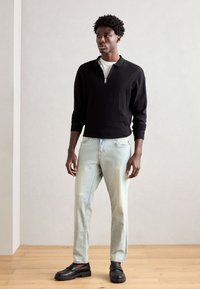 Black zippered pullover, cotton blend, long sleeves. Light blue straight-leg jeans. Black shoes with a chunky sole. Minimalist backdrop.