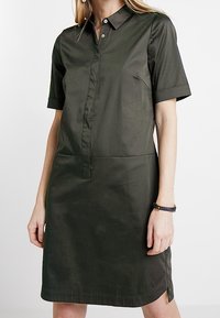 Sukienka z krótkim rękawem w ciemnozielonym kolorze, wykonana z gładkiej tkaniny. Posiada kołnierzyk, zapinana na guziki z przodu oraz ma boczne rozcięcia, które ułatwiają ruch.