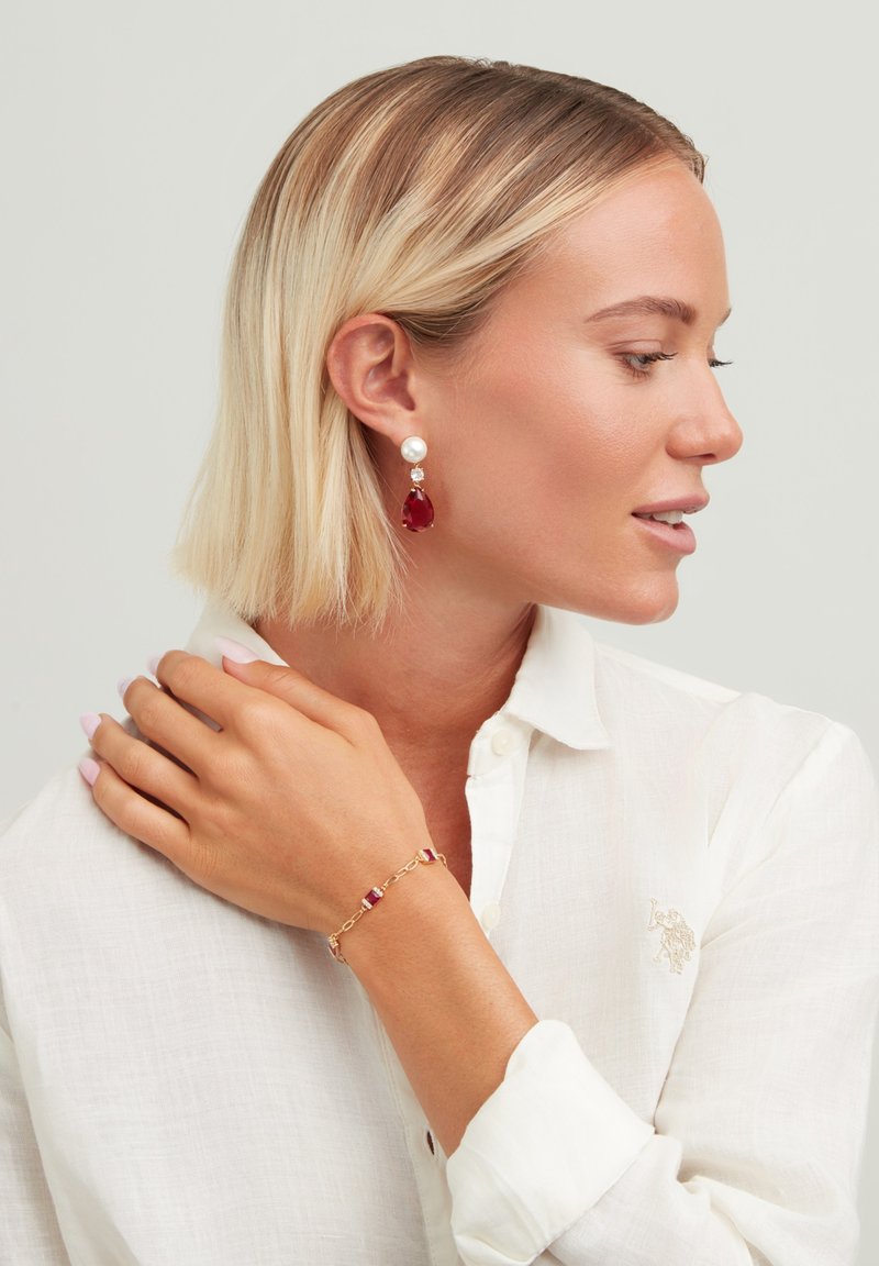 Bracelet en or avec des accents rouges ; boucles d'oreilles imposantes avec une perle et une pierre précieuse rouge ; porté avec une blouse blanche.