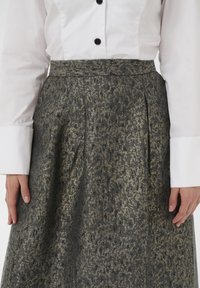 Knee ilgio sijonas, pagamintas iš struktūrinio audinio su juodu ir auksiniu raštu, su klostėmis ir prigludusia juosmena, derinamas su balta marškinėlių palaidine.