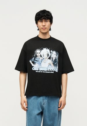 Young man wearing a black oversized T-shirt with a graphic of three women and text promoting voting for favorite artists.