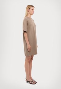Svetlohnedé oversized tričko-dress s krátkymi rukávmi, okrúhlym výstrihom a hladkým povrchom, v kombinácii s čiernymi sandálmi na podpätku s popruhmi.