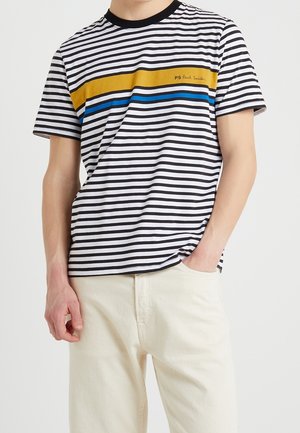 Homme portant un T-shirt rayé noir et blanc avec des bandes horizontales jaunes et bleues et un pantalon beige clair, main dans la poche.