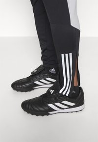 Schwarze Sportschuhe mit weißen Akzenten, einer strukturierten Oberseite und drei weißen Streifen. Kombiniert mit schwarzen Hosen, die graue Abschnitte und Reißverschlüsse aufweisen.