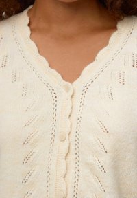 Cardigan tricoté de couleur crème avec un décolleté festonné, une fermeture à boutons et de magnifiques motifs de feuilles sur le devant. Texture douce.