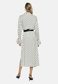 Vestito a maniche lunghe bianco con pois neri, dotato di una vita con cintura, polsini arricciati e spacco laterale per facilitare il movimento.