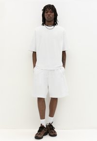 T-shirt blanche oversize à manches courtes et shorts assortis en tissu peluche. Baskets marron avec accents noirs, colliers superposés et chaussettes blanches.