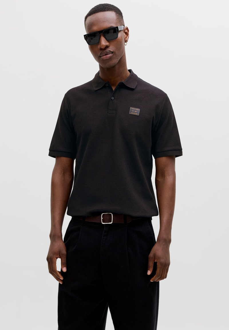 Homme portant un polo noir, un pantalon noir, une ceinture marron, des lunettes de soleil noires et des boucles d'oreilles en anneaux dorés, debout devant un fond blanc.