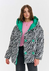 Žena oblečená v čierno-bielom pufrom s pruhmi zebra a zeleným podšívkovým látkom, ružovom svetri, čiernych nohaviciach a zlatom náhrdelníku.