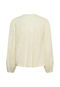Blouse crème à manches longues avec texture à pois, coupe ample et poignets élastiques froncés, vue de dos sur fond blanc.