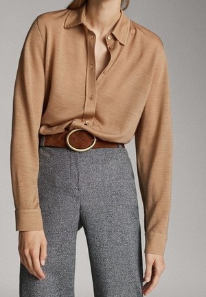 Personne portant une chemise camel à manches longues boutonnée rentrée dans un pantalon taille haute gris avec une ceinture marron comportant une grande boucle circulaire.