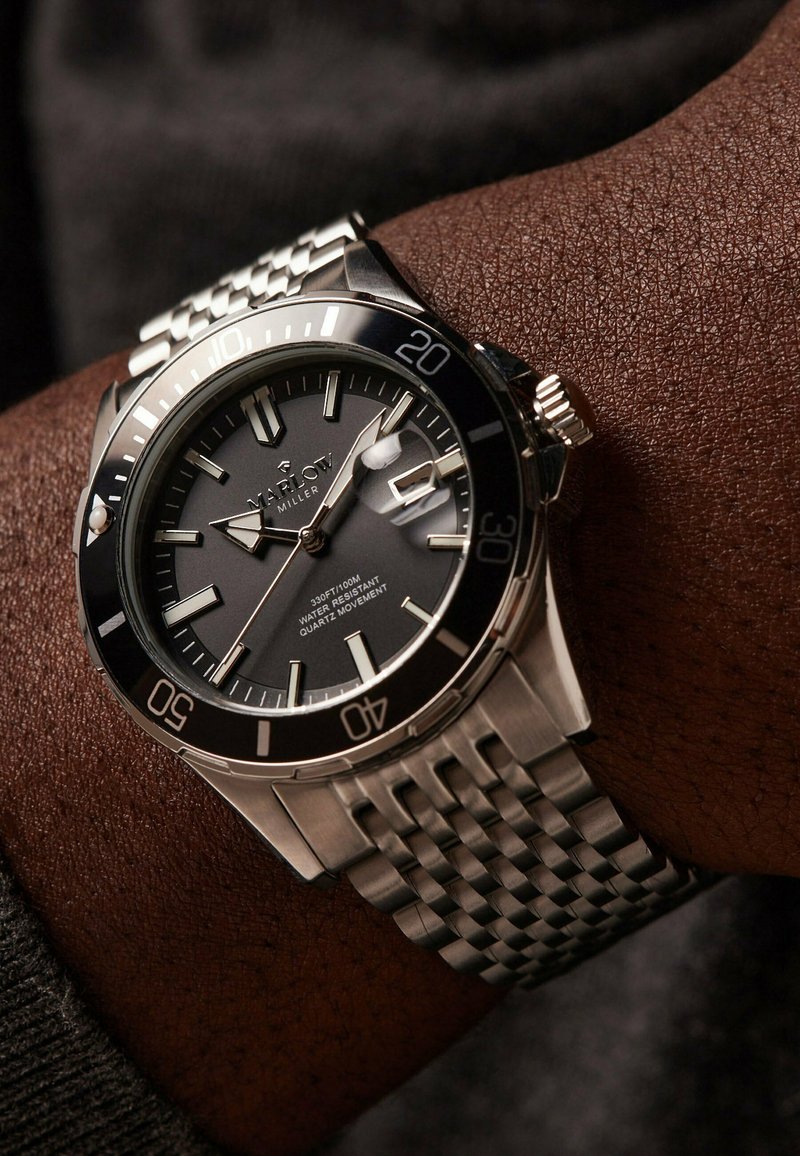 Silver stainless steel watch with a black dial, luminous markers, date window, and a textured metal bracelet. Water-resistant up to 100m.