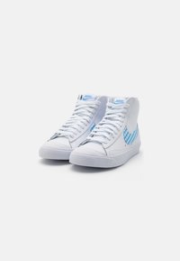 Par belih Nike visokih superg z modrim gingham vzorcem na swooshu in svetlo modrim Nike logotipom na jeziku.