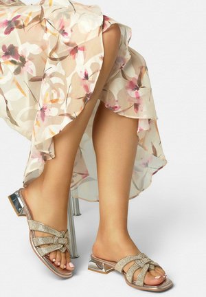 Vrouw draagt een doorschijnende jurk met bloemenprint en zilverkleurige open-toe sandalen met lage hak en strikaccenten van strass-steentjes, zittend met gekruiste benen.