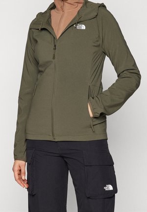 Person iført en grøn lynlåstrøje og sorte cargobukser, begge med The North Face-logo, stående med den ene hånd i lommen.