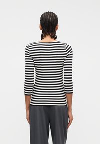 Άτομο με κοτσίδες που φοράει τοπ με λευκές και μαύρες οριζόντιες ρίγες και μανίκια τριών τετάρτων και σκούρο γκρι παντελόνι, γυρισμένο με την πλάτη.