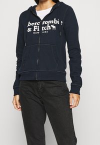 Felpa con zip di colore navy realizzata in tessuto morbido, dotata di una tasca a canguro frontale, cappuccio con coulisse e testo del logo bianco sul petto.