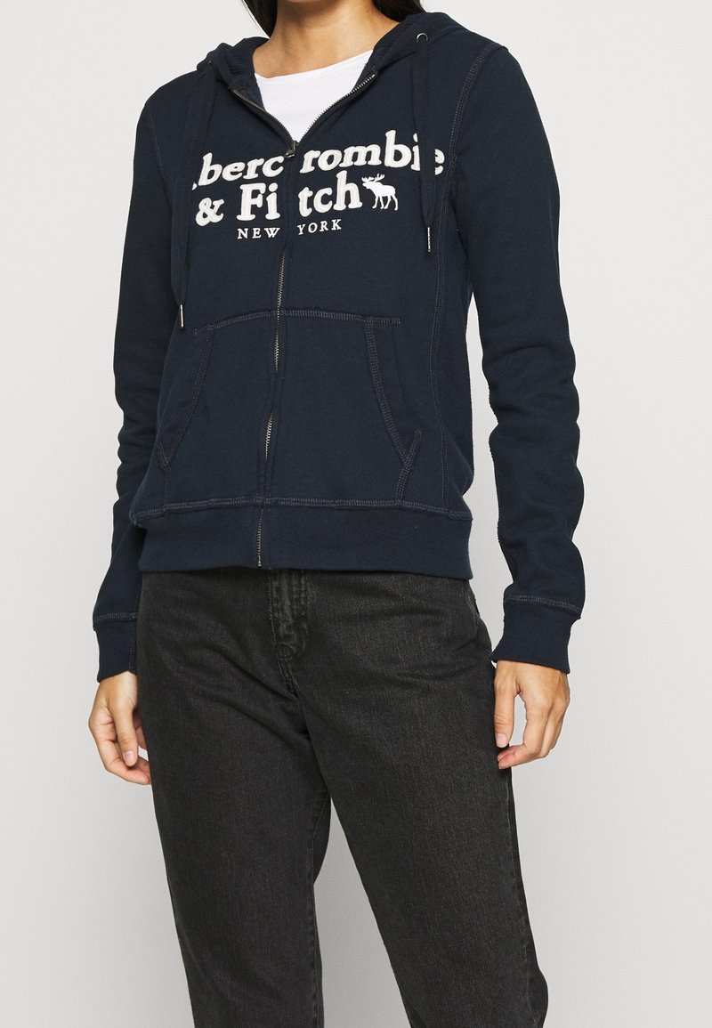 Felpa con zip di colore navy realizzata in tessuto morbido, dotata di una tasca a canguro frontale, cappuccio con coulisse e testo del logo bianco sul petto.