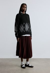 Maglione nero lavorato a maglia con motivo argyle bianco e grigio, abbinato a una gonna plissettata color marsala, camicia bianca con colletto e scarpe nere con calzini bianchi.