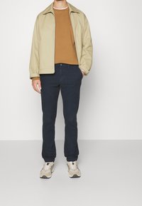 Giacca in nylon khaki sopra un maglione marrone, pantaloni blu navy e sneakers beige. La giacca ha un colletto classico e chiusura con zip; i pantaloni presentano un motivo testurizzato.