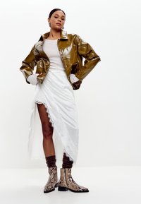 Chaqueta de vinilo brillante color verde oliva, falda envolvente blanca con borde de encaje y calcetas marrones con textura de canalé. Botines de serpiente en el tobillo completan el look.