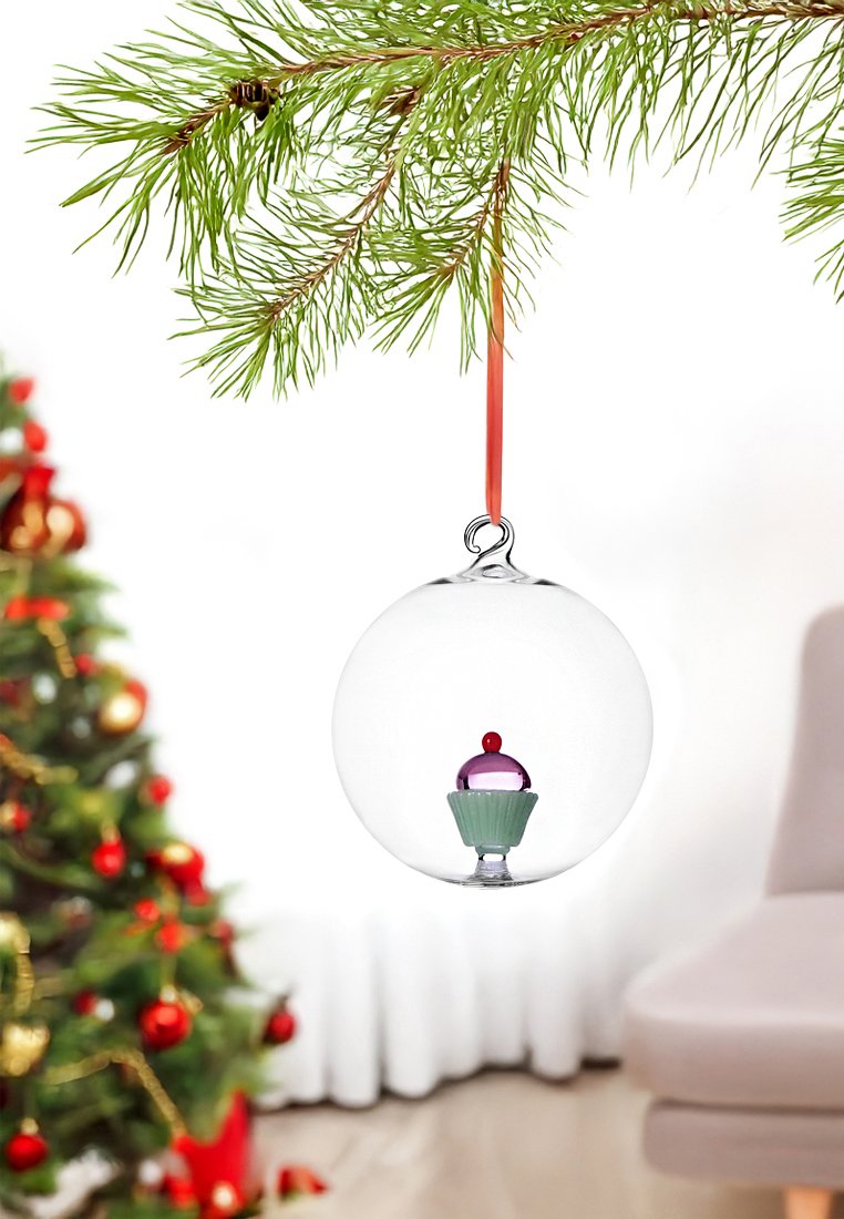 Przezroczysta szklana ozdoba z różowym i fioletowym babeczką w środku, zawieszona na czerwonej wstążce, wisząca na zielonej gałęzi sosny.