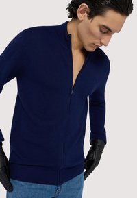 Marinblå zip-up tröja med en texturerad stickad design, ribbade manschetter och fåll. Bärs med svarta handskar, vilket visar en modern, åtsittande silhuett.