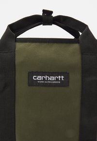 Oliwkowa zielona torba materiałowa z czarnymi uchwytami. Posiada czarną etykietę z białym napisem "carhartt WORK IN PROGRESS." Solidne szwy i minimalistyczny design.