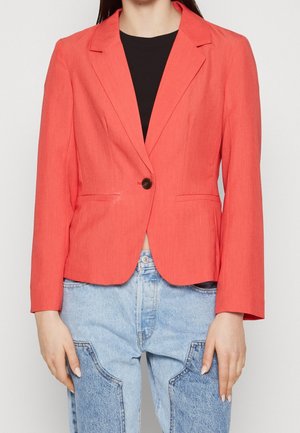 Kvinde iført en koralfarvet blazer med enkelt knap over en sort top og lyseblå jeans med lapper, stående foran en ensfarvet baggrund.