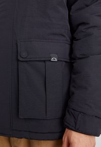 Marineblaue Steppjacke mit einer Klappentasche mit Knopfverschluss, strukturiertem Stoff und elastischen Bündchen für eine enganliegende Passform.