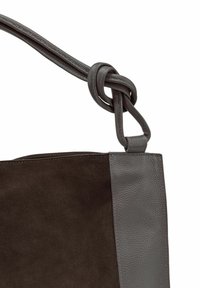 Sac en cuir avec un corps en daim marron foncé et un panneau latéral en cuir gris contrastant. La sangle présente un détail noué pour un design supplémentaire.