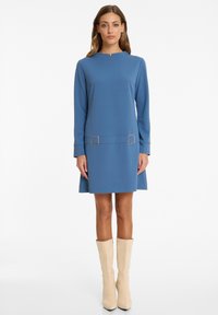 Blaues, langärmliges Kleid mit höherem Ausschnitt und zwei goldenen quadratischen Akzenten in der Taille. Glatte Textur, knielang, taillierte Passform.