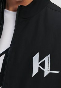 Svart jacka med hög krage, som har en silverlogotyp på vänster bröst och en hel dragkedja. Slät, lättviktsmaterial.