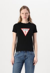 T-shirt en coton noir avec un logo triangulaire rouge audacieux portant le texte "GUESS ?". Manches courtes et coupe classique. Un jean en denim bleu complète le look.